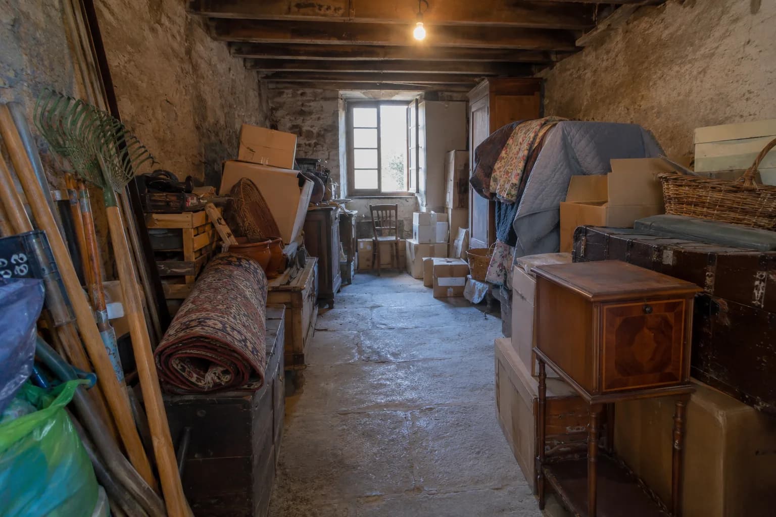 Cave encombrée de meubles et de cartons empilés le long des murs, avec un passage dégagé au centre